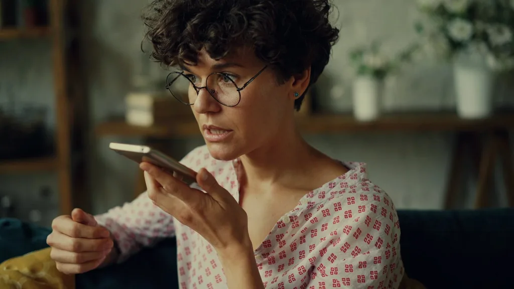 A woman in round glasses holds her phone close to her mouth to record a voice message at home.