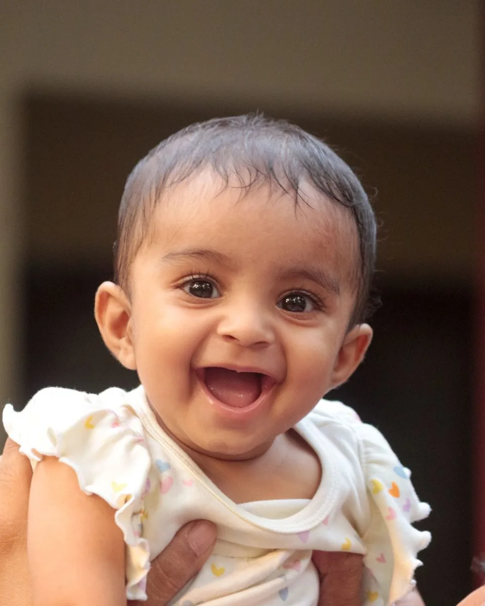 Laughing baby girl in a heart-print top, held up by a parent's hands.