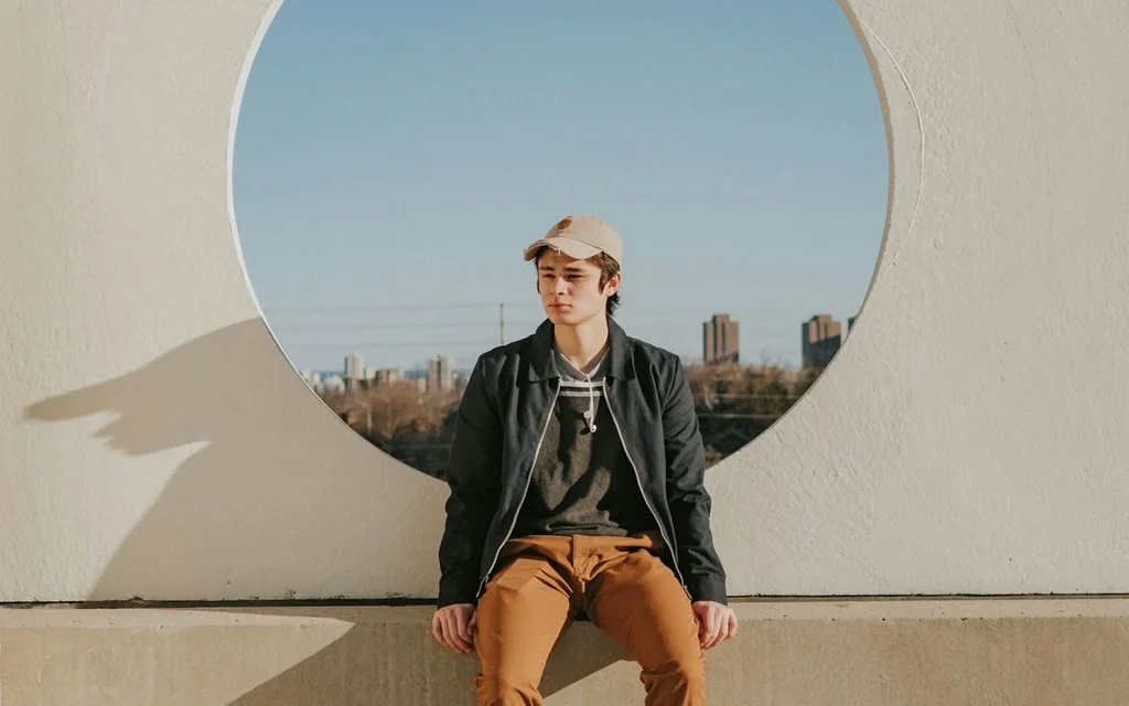 Young man in a cap sitting below a round window cut into a white wall.