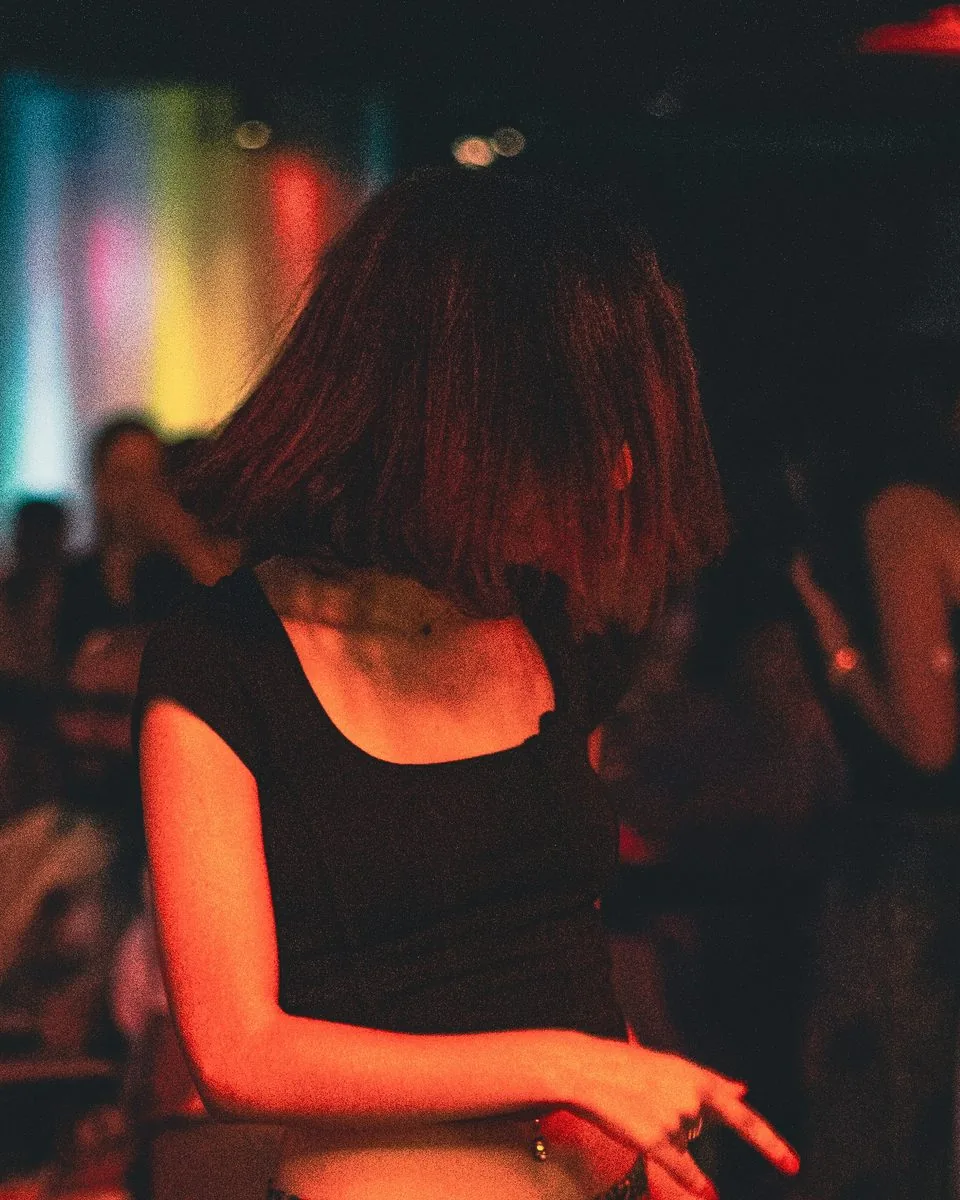 Young woman dances in a dim nightclub, hair swinging under warm red and rainbow lights.