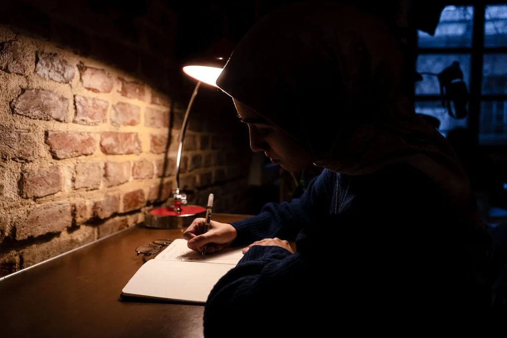 A woman writes in a notebook under a desk lamp in a dark room with a brick wall.