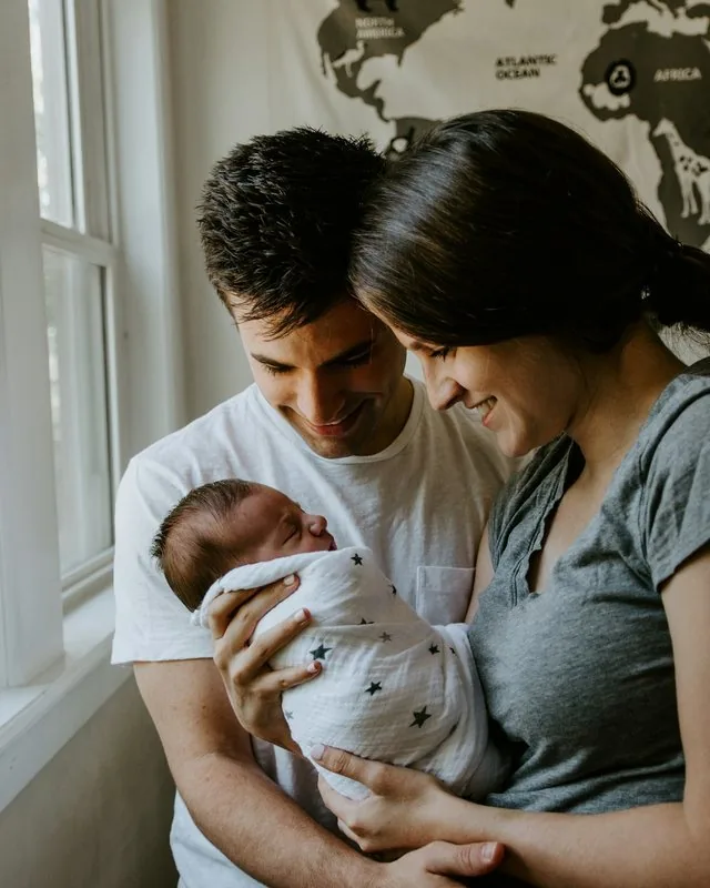 Young couple smiles down at their sleeping newborn beside a bright window.