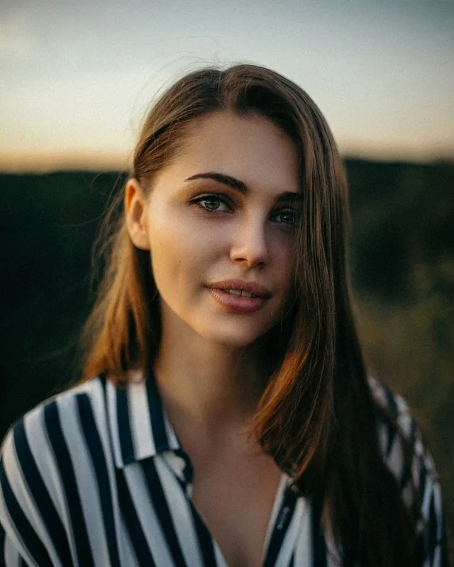 Young woman in a striped shirt smiles softly in golden evening light.