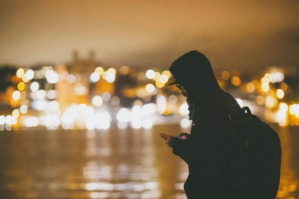 Hooded man with a backpack checks his phone, silhouetted against blurred gold city lights on water.