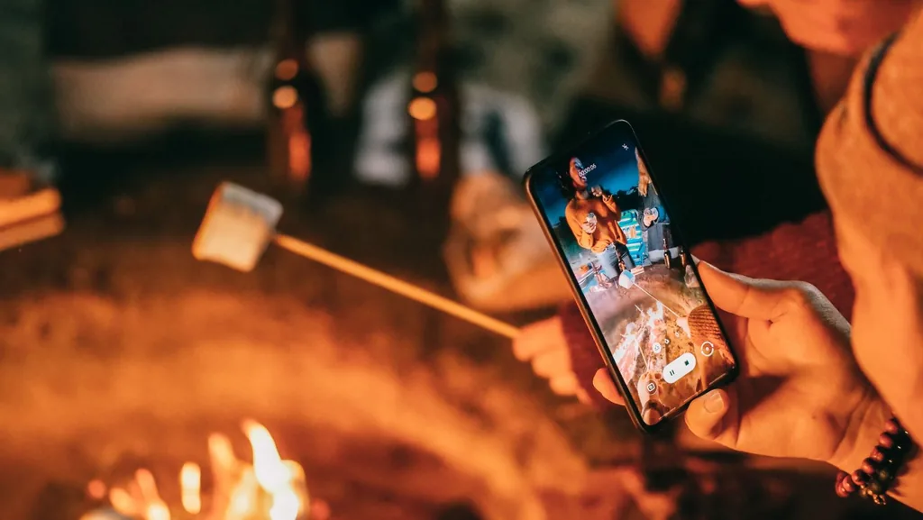 A phone records friends roasting marshmallows around a glowing campfire at night.